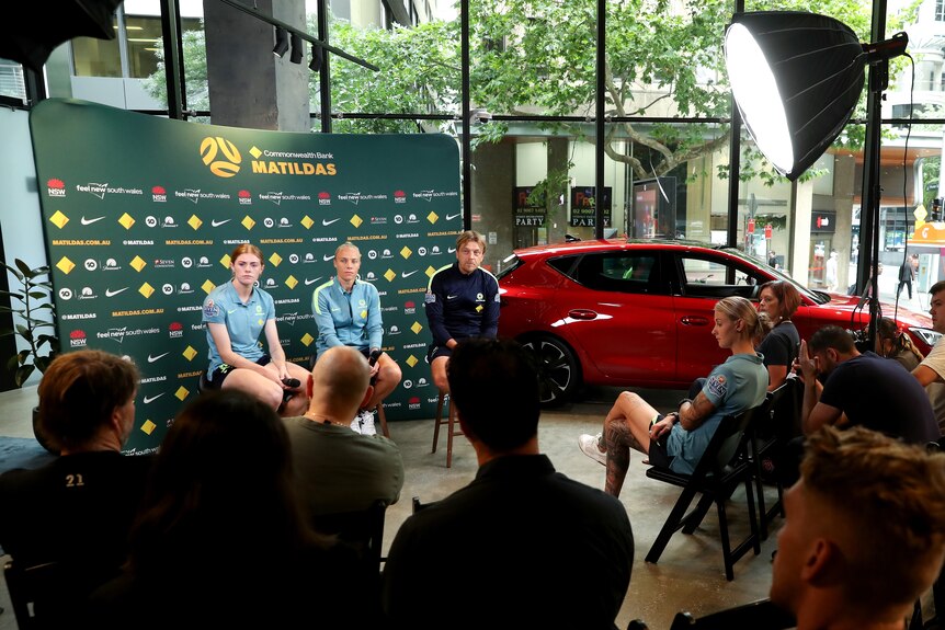 Two soccer players and a coach sit on some chairs in front of an advertising screen as they address the media in the room