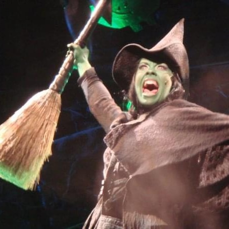 Erin Cornell performs onstage as Elphaba. She is wearing a witch's hat and cape, and has green skin.