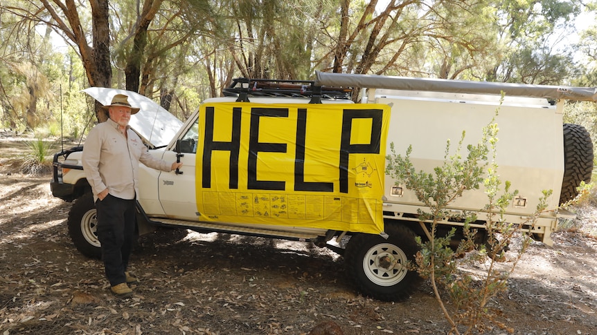 A man stands beside a broken-down vehicle with a big yellow sign on the side saying 'help' in black letters.