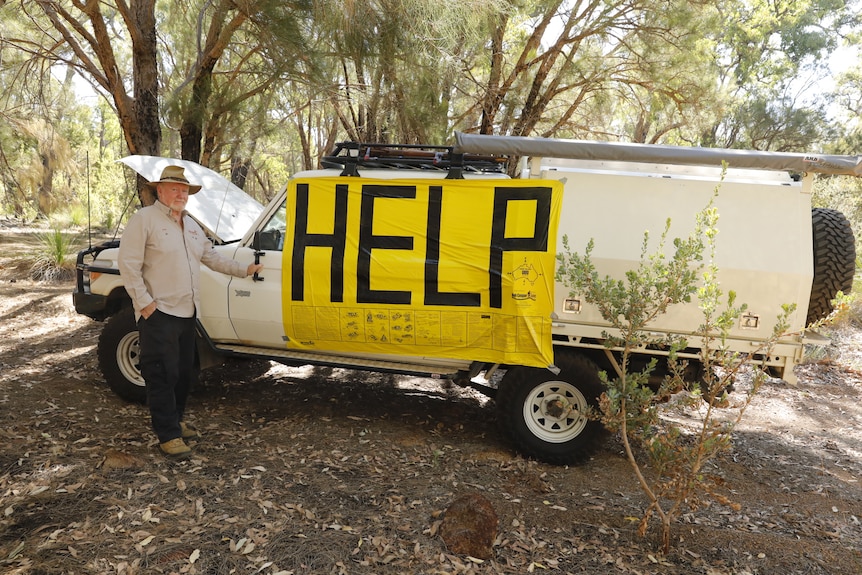 A man stands beside a broken-down vehicle with a big yellow sign on the side saying 'help' in black letters.