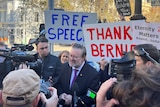 A man in a suit and tie is photographed and speaks to reporters, while people hold up supportive signs.