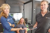 A man and a woman hold out small technical equipment while another man sits on a squat rack.
