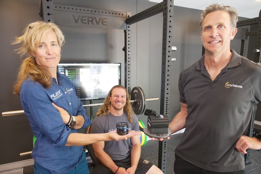 A man and a woman hold out small technical equipment while another man sits on a squat rack.