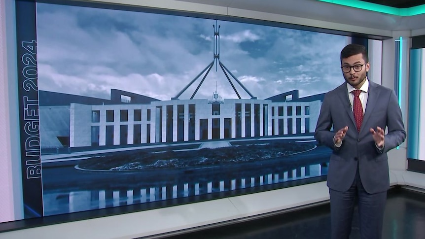 A man speaks in a television studio with a large graphic of Parliament House behind him.