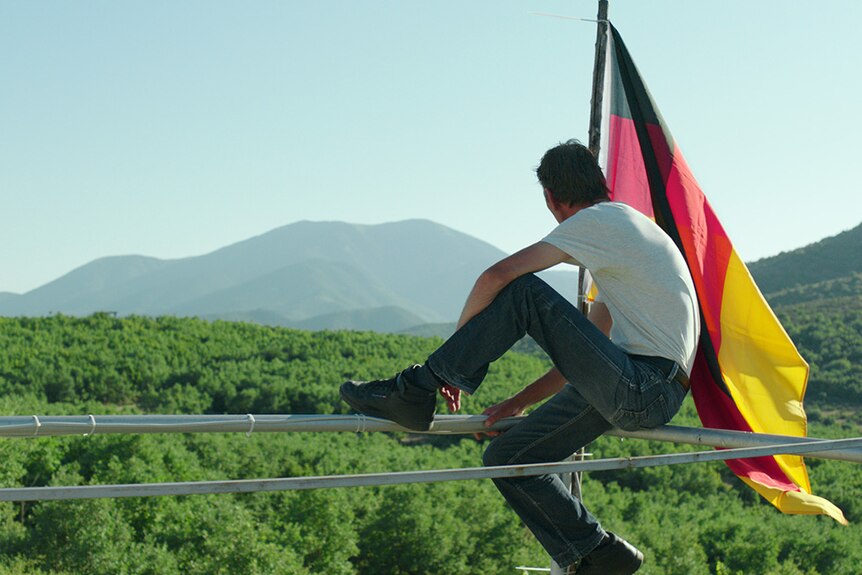 A man holding a large German flag sits perched on a fence looking out over a green valley.