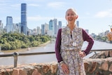 Judi Adams stands wearing purple long sleeve top under a paisley print dress with Brisbane river and brisbane city in background