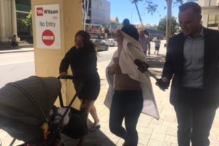 Two people walk along the footpath covering their faces, with one pushing a pram and a reporter walking along side.