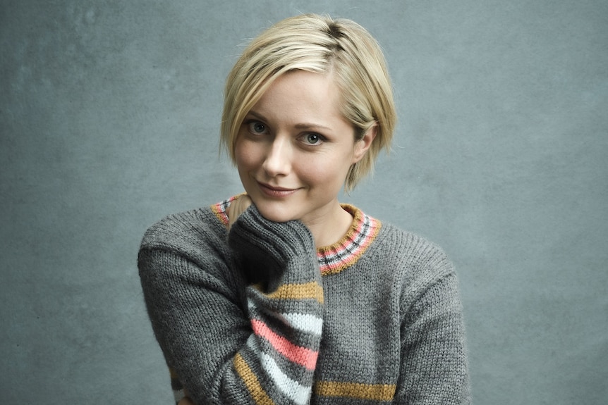 Georgina Haig sits on a stool in a jumper and jeans, smiling.