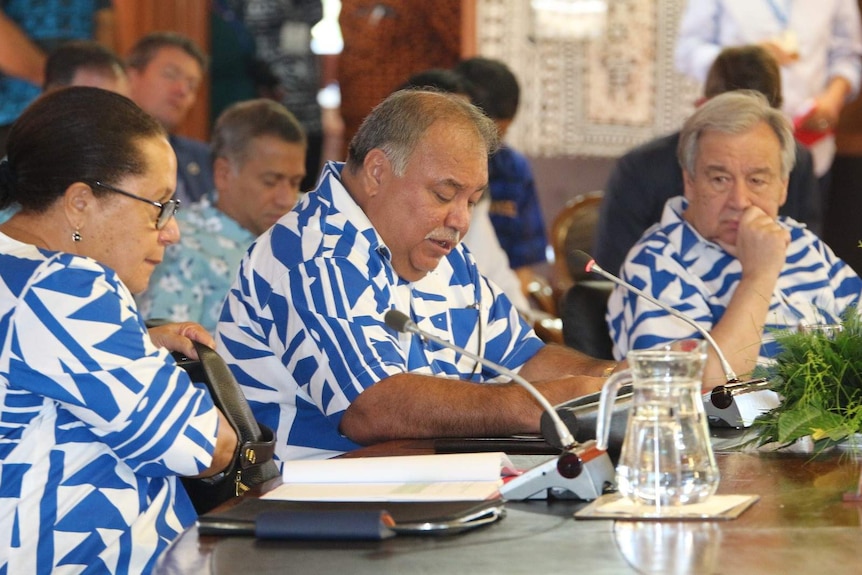 Three people wearing matching blue and white shirts sit at a conferences table speaking into microphones.