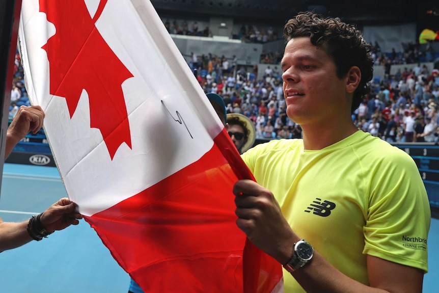 A smiling tennis player holds a Canadian flag as he signs it with a marker.