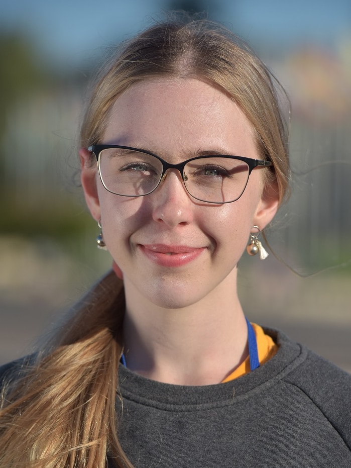 Portrait shot of a young woman with blonde hair, glasses and a grey jumper smiling.