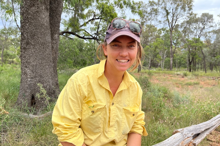 A young woman in yellow shirt, pink cap, goggles on tip, sits in a park, trees behind.