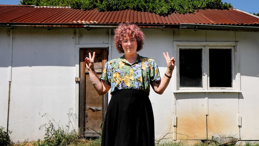 A trans person with red curly hair, a long black skirt and colourful blouse stands in front of a shed doing the "peace sign"