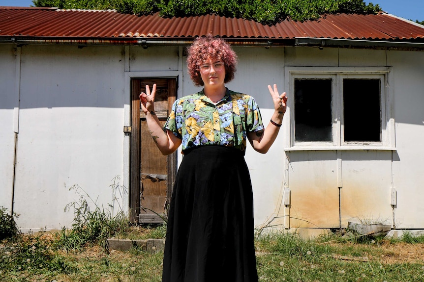 A trans person with red curly hair, a long black skirt and colourful blouse stands in front of a shed doing the "peace sign"