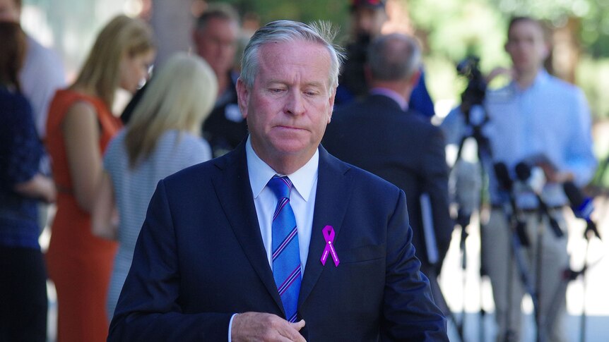 Colin Barnett looks downcast as he walks away from journalists, wearing a pink ribbon on his jacket.