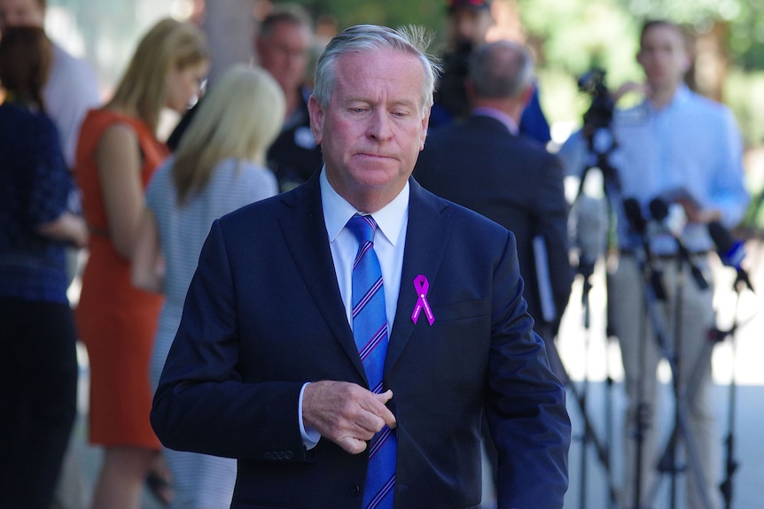 Colin Barnett looks downcast as he walks away from journalists, wearing a pink ribbon on his jacket.