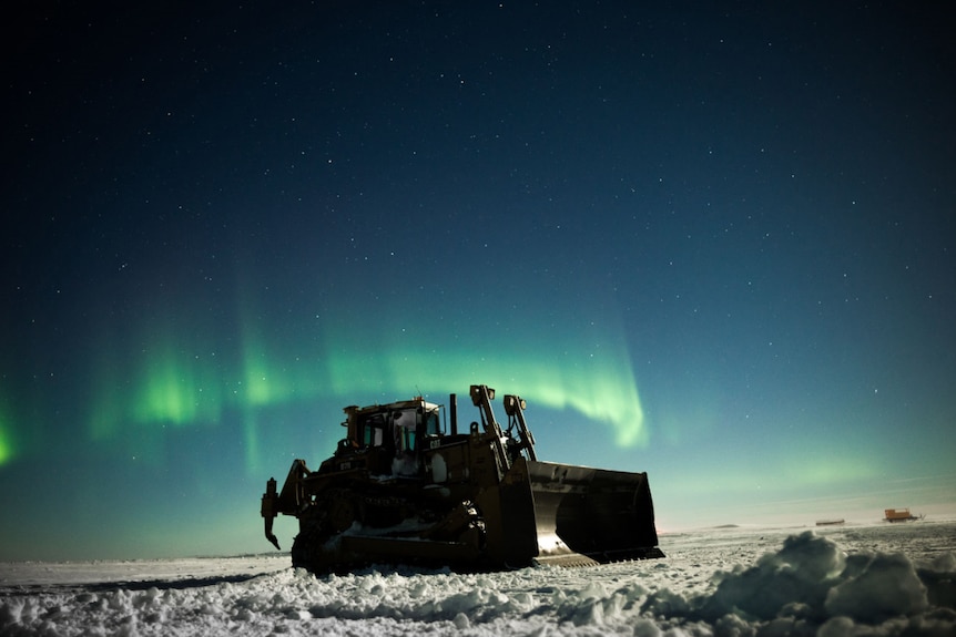 A snow grader at night, with an Aurora Australis in the night sky.