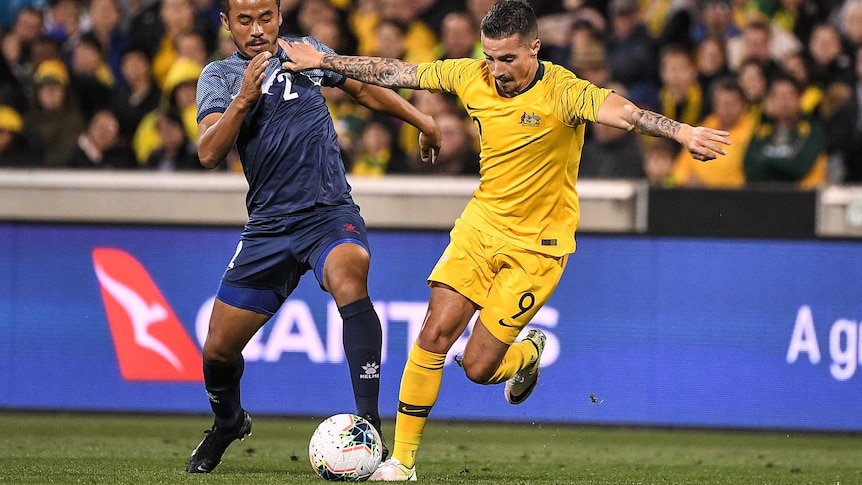 Jamie Maclaren, wearing gold football kit, runs with the ball at his feet next to a player wearing a blue shirt