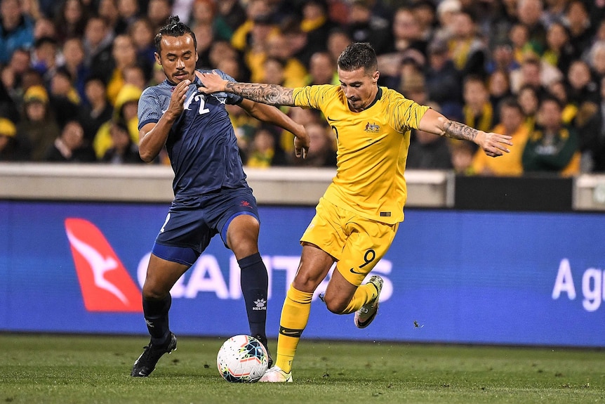 Jamie Maclaren, wearing gold football kit, runs with the ball at his feet next to a player wearing a blue shirt