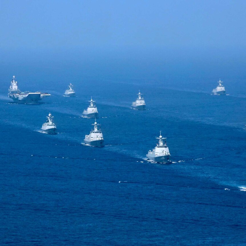 An aircraft carrier is accompanied by navy frigates and submarines in the South China Sea.
