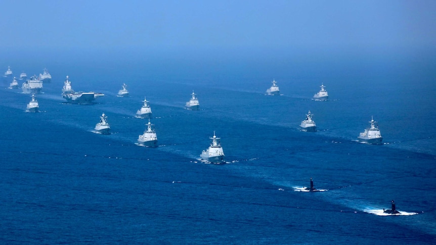 An aircraft carrier is accompanied by navy frigates and submarines in the South China Sea.