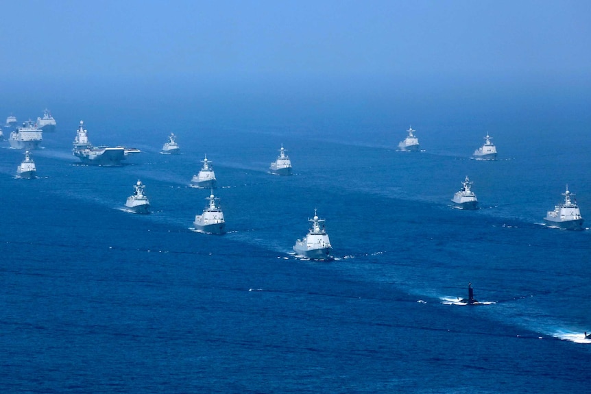 An aircraft carrier is accompanied by navy frigates and submarines in the South China Sea.