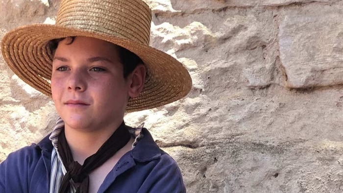 A young boy in a straw hat stands against a sandstone wall