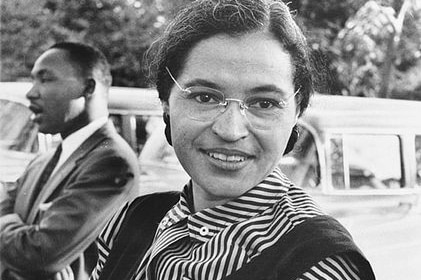 Rosa Parks leans against a car and smiles with Martin Luther King Jr in the background.
