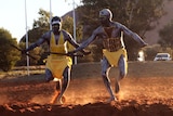 Two men wearing yellow white and brown body paint and a yellow outfit dance in front of Uluru.