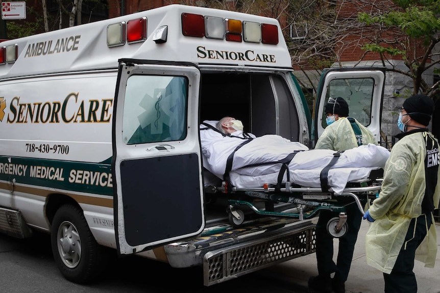 An older person with grey hair is loaded into an ambulance in a stretcher by medical workers wearing masks.