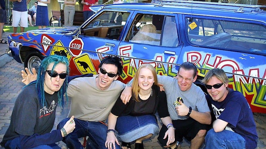 A young woman in a denim skirt kneels in front of a Holden Kingswood with unkempt men surrounding her.