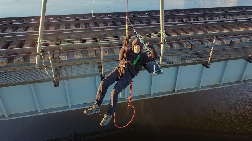 A man hanging from a rope above a train line.