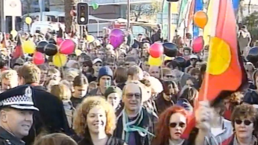 Crowd marches in street, wave Aboriginal flag