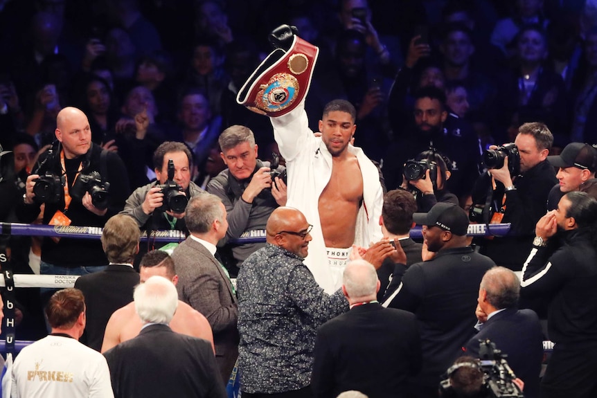 Boxer Anthony Joshua raises a belt surrounded by photographers in the ring