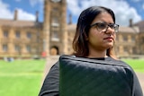 A young woman of Indian descent stands in the grounds of Sydney University wearing a black top and is holding her laptop.