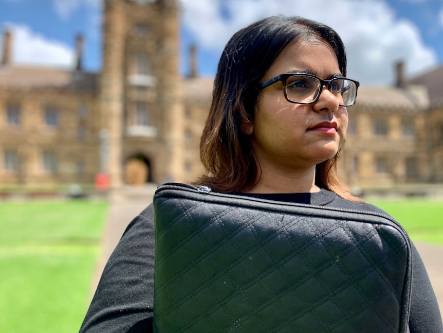 A young woman of Indian descent stands in the grounds of Sydney University wearing a black top and is holding her laptop.