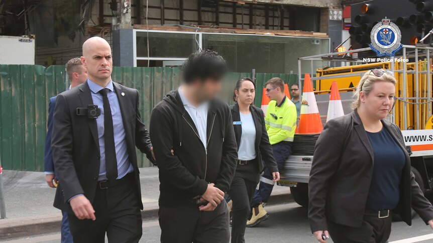 NSW detectives escort a male suspect, whose face is blurred, next to a parked construction ute.