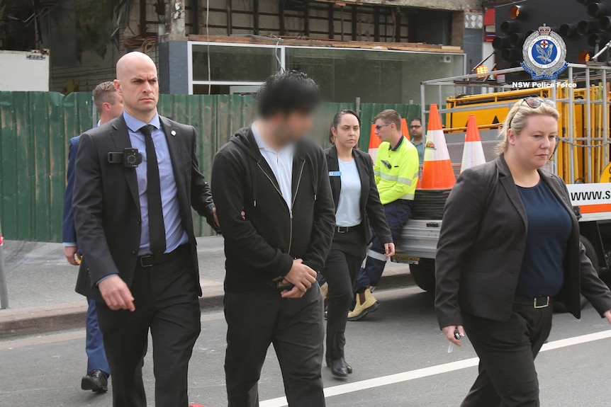 NSW detectives escort a male suspect, whose face is blurred, next to a parked construction ute.