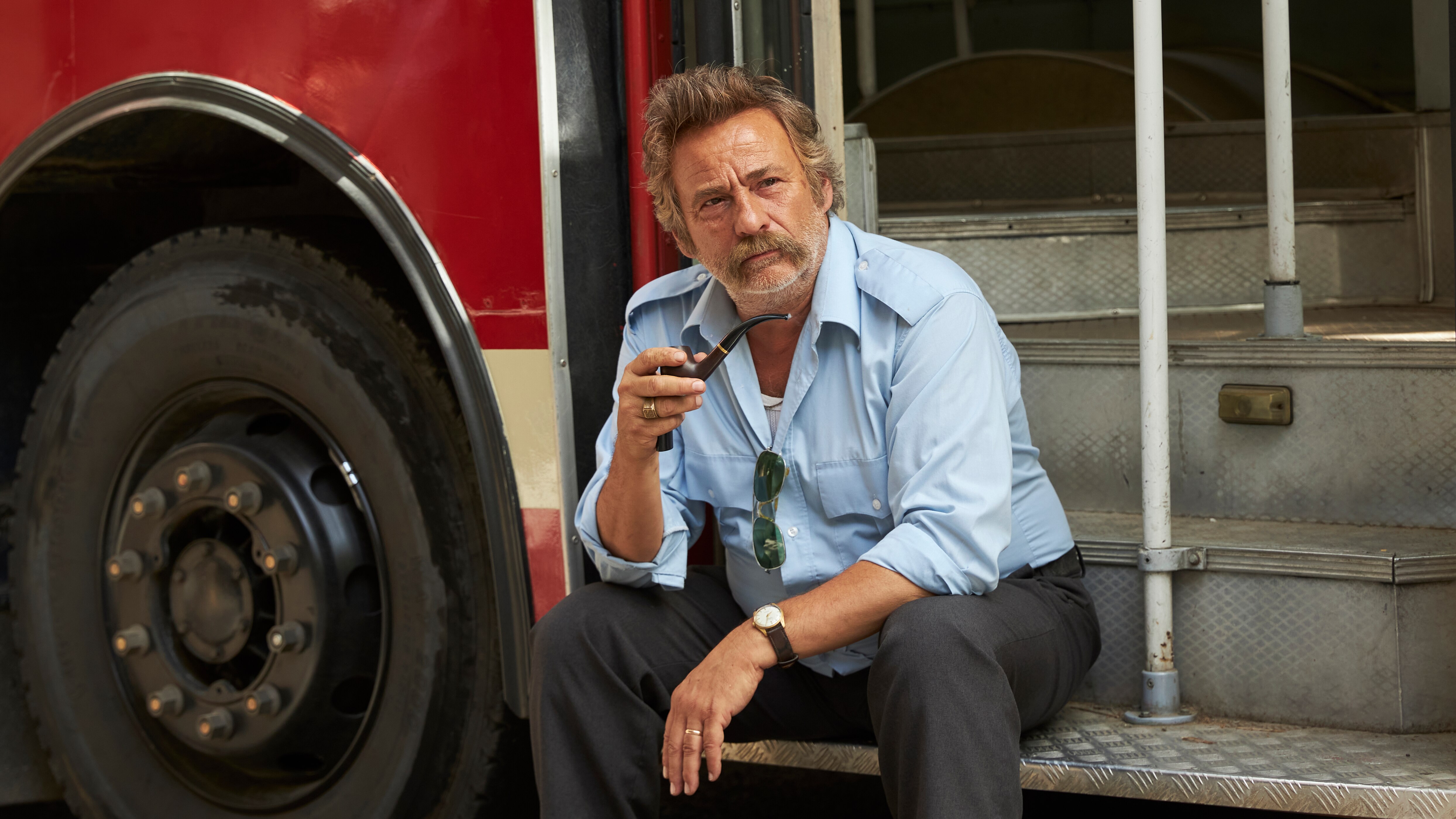 A man dressed as a bus driver sits on the steps of a bus while holding a smoke pipe in his right hand.