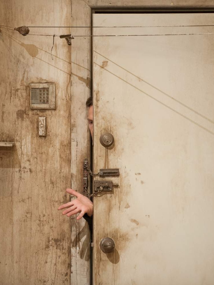 An actor reaches out from behind a dirty, chain-locked door in the play Give Me Your Love.