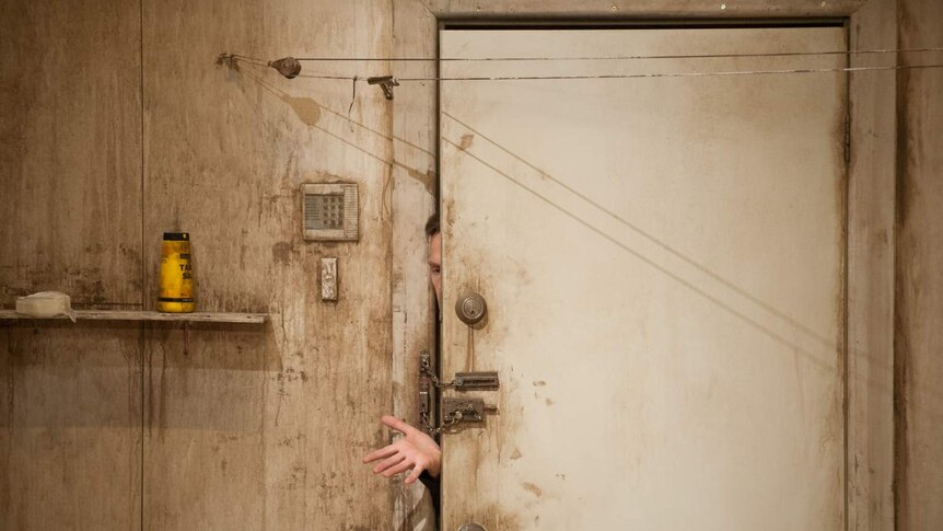 An actor reaches out from behind a dirty, chain-locked door in the play Give Me Your Love.
