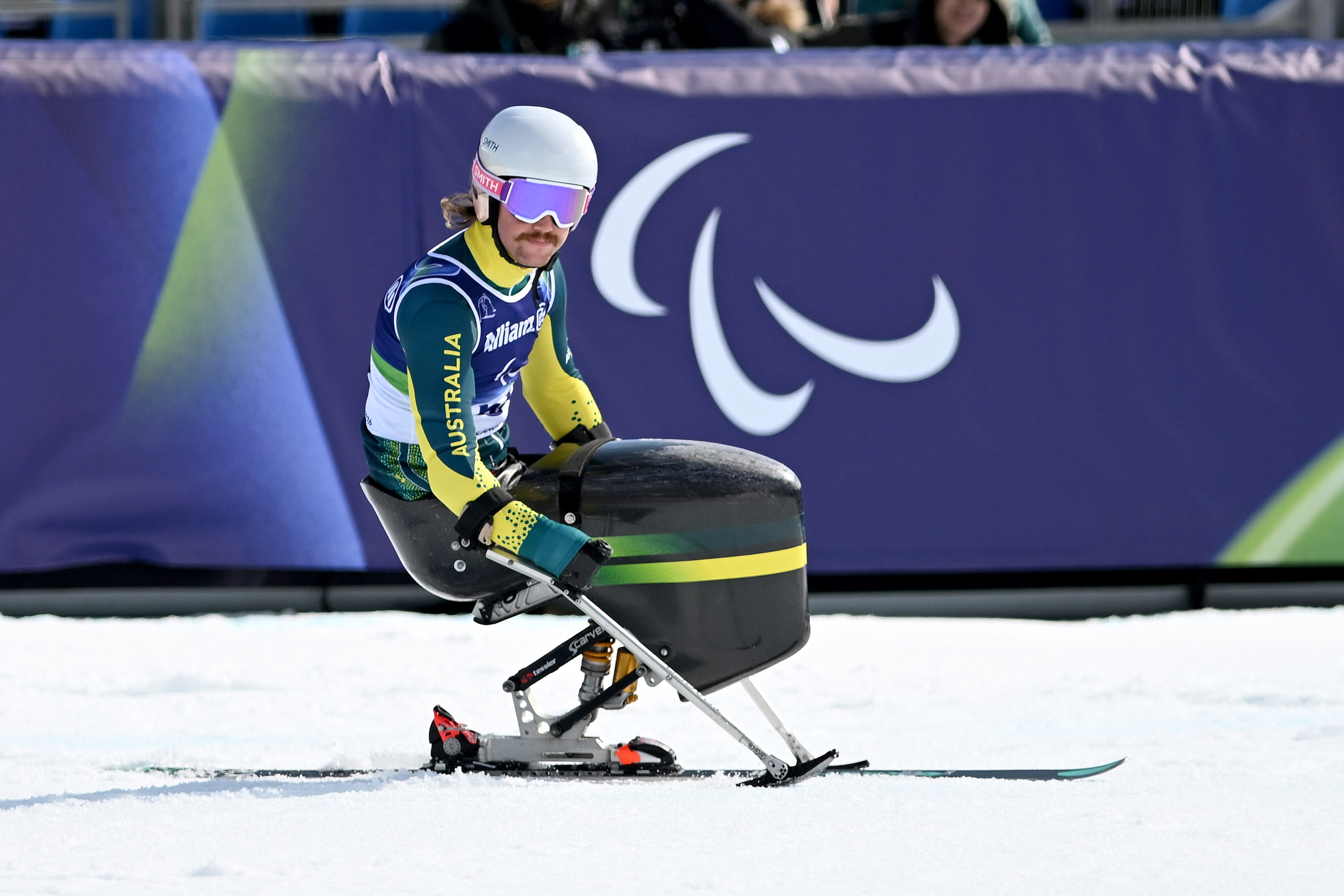 Um esquiador sentado, vestindo verde e dourado, está sentado na neve esperando para ir.