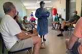 People sit in waiting room for a covid vaccine while a healthcare worker wearing PPE stands in the room.