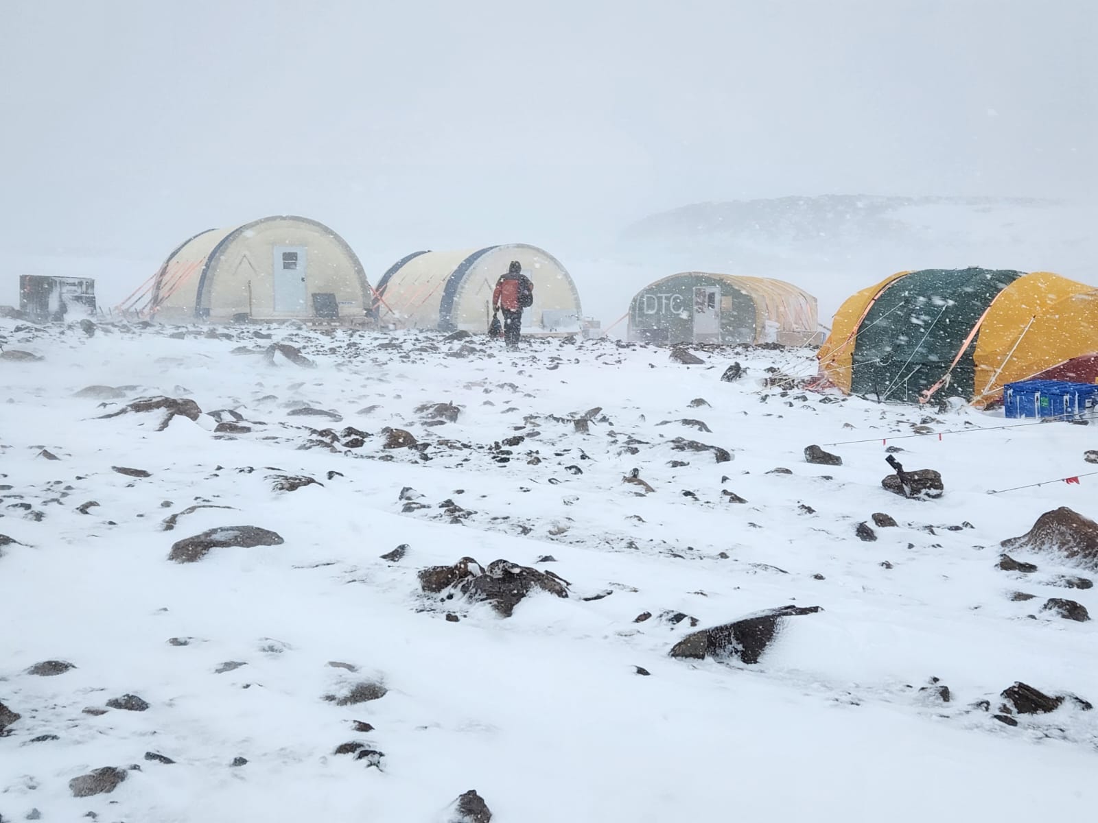 A man in winter gear walks through a blizzard toward three yellow tents.