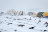 A man in winter gear walks through a blizzard toward three yellow tents.