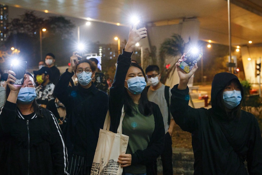 An Asian woman in a green top and face mask holds a lit mobile phone in a group in a city setting.