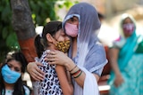 A woman wearing a hijab and a mask holds onto a young girl crying while wearing a mask