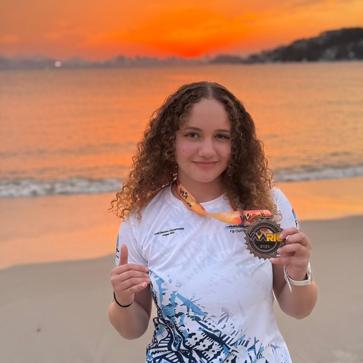 A young Fijian girl stands on the beach with an orange sunset in the background holding a bronze medal around her neck.
