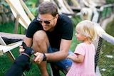 A smiling man wearing black glasses crouches in front of a small black dog while a young girl looks on beside him.