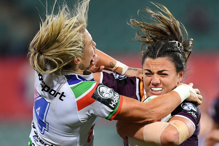 A Brisbane Broncos NRLW player holds the ball as she attempts to palm off a Warriors opponent.